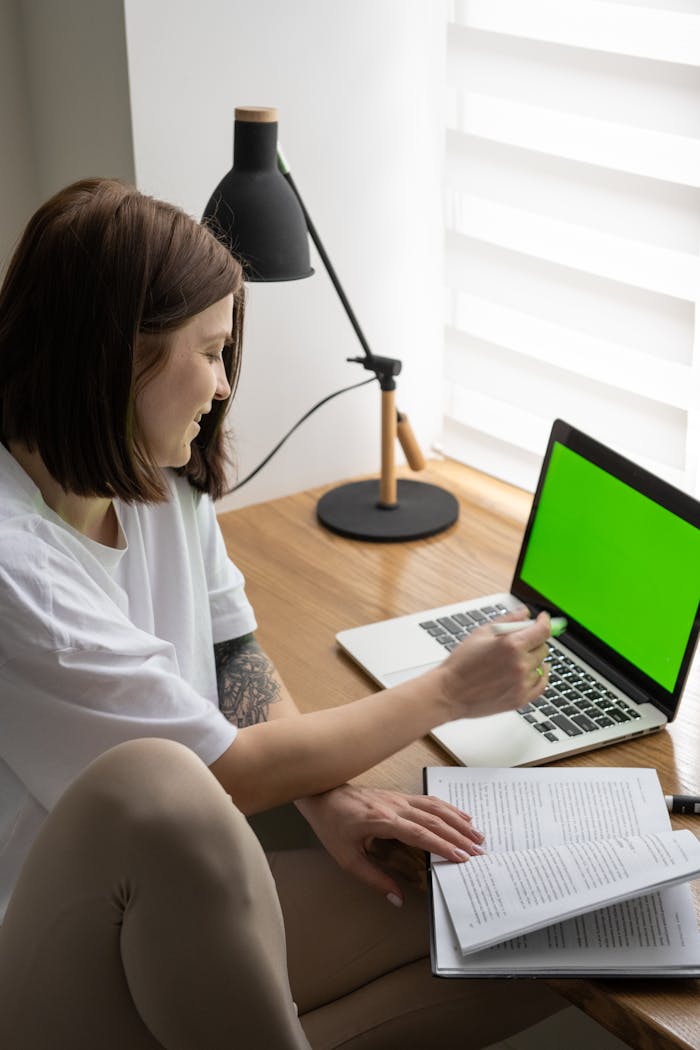 From above side view of cheerful female student sitting at table with laptop with chromakey screen and reading book while doing homework assignment